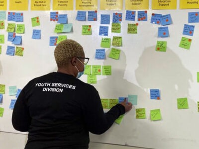 A Black woman with short bleached hair and glasses places a sticky note on a whiteboard.