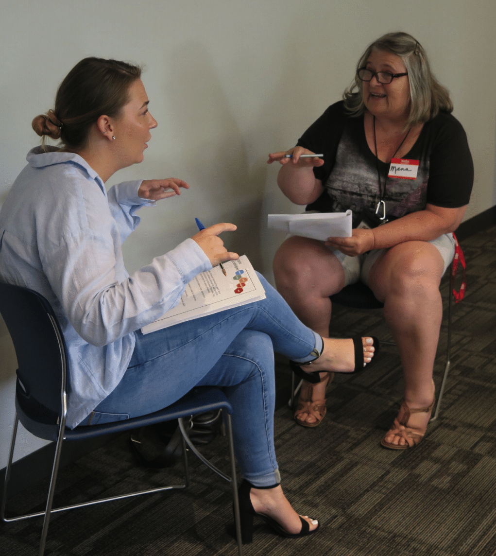 Two women sitting on chairs with notebooks talking to each other.