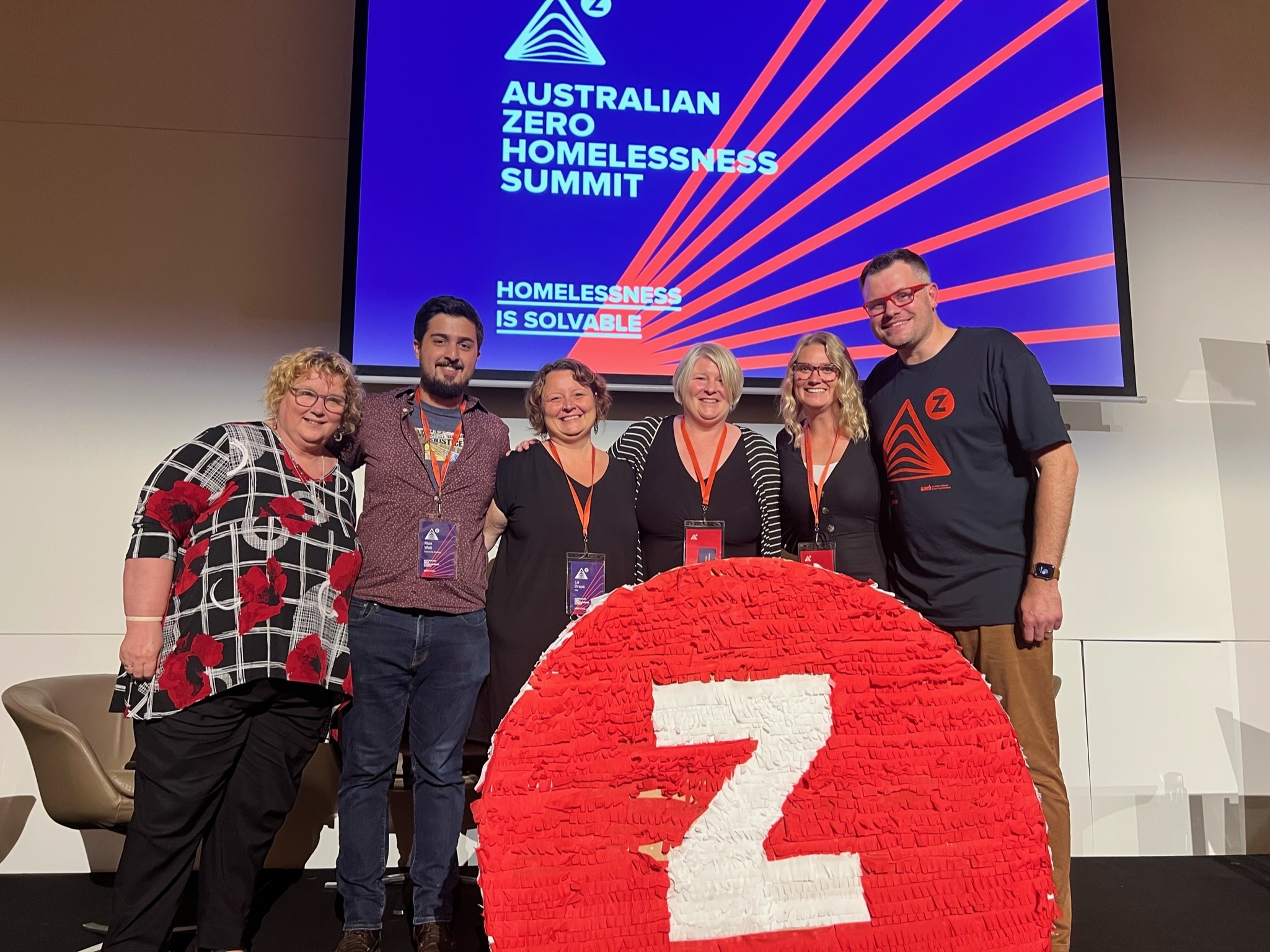 Group photo of six people standing on a stage in front of a large Z