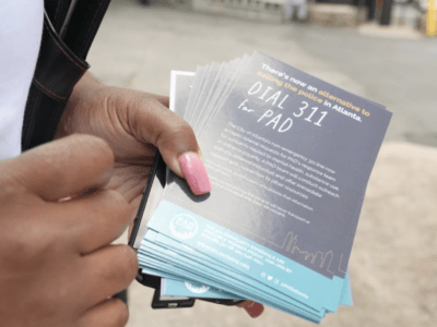 Image of a hand holding small flyers saying "There's now an alternative to calling the police in Atlanta. Dial 311 for PAD."
